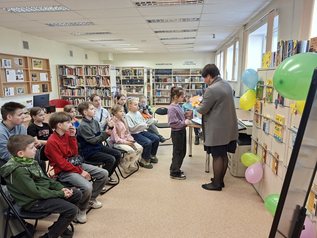 Bibliotekāre bibliotēkā pasniedz diplomu mazai meitenei. Rindās sēdošie bērni uzmanīgi vēro notiekošo. Telpa ir dekorēta ar krāsainiem baloniem, un plauktos sarindotas grāmatas.