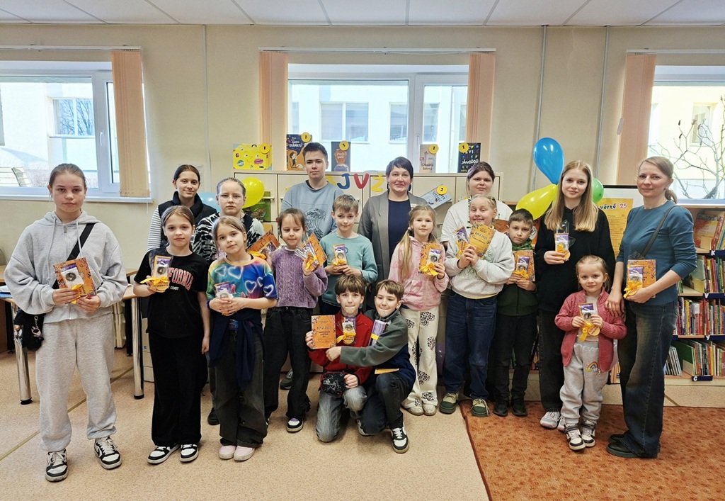 Smaidīgu bērnu un pieaugušo grupa bibliotēkā, tur rokās grāmatas un kartītes. Telpa ir dekorēta ar baloniem un košiem plakātiem. Fonā redzamas grāmatas un grāmatu plaukts labajā pusē.