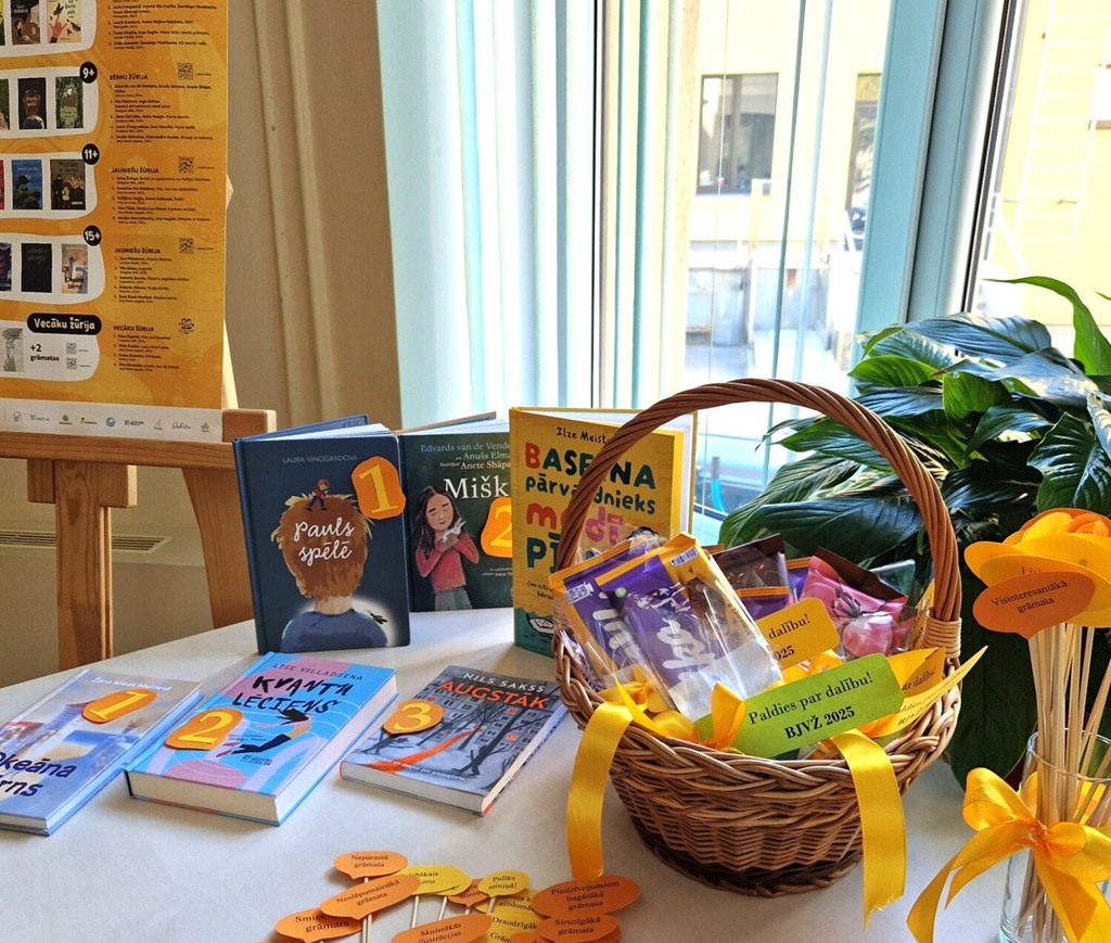 Uz galda izstādītas grāmatas ar numurētām norādēm. Blakus atrodas grozs ar krāsainiem kārumiem un dzeltenām lentēm. Fonā pie gaiša loga redzams plakāts un telpaugs.