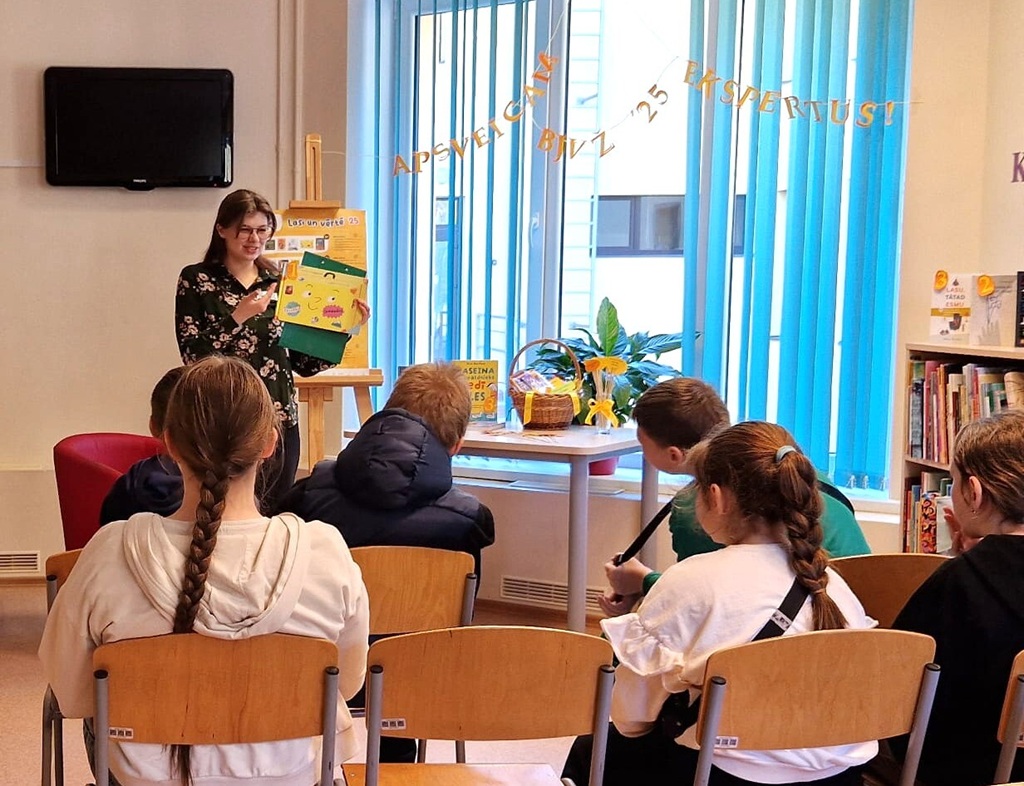 Sieviete stāv bibliotēkas priekšā ar krāsainu grāmatu rokās un prezentē to krēslos sēdošiem bērniem. Telpa ir gaiša un dekorēta, radot dzīvīgu un saistošu atmosfēru.