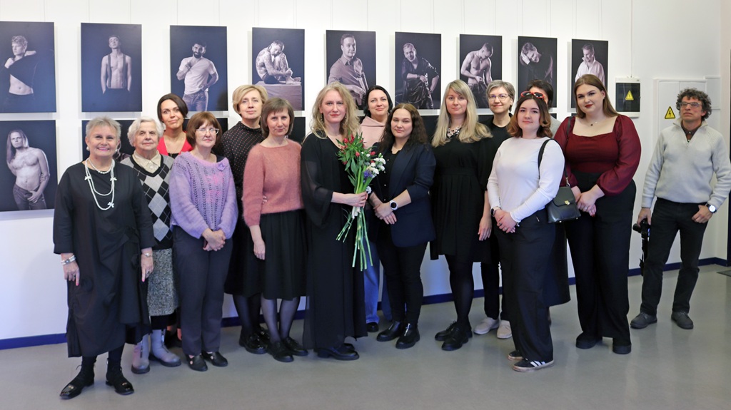Sieviešu grupa stāv kopā pie sienas, uz kuras ir izvietoti dažādi attēli.