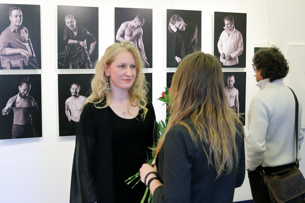 Sieviete sarunājas ar citu sievieti pie sienas, uz kuras ir izvietotas dažādas melnbaltas fotogrāfijas.