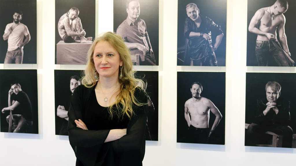 Sieviete stāv ar sakrustotām rokām pie sienas, uz kuras redzami melnbalti vīriešu portreti dažādās pozās.