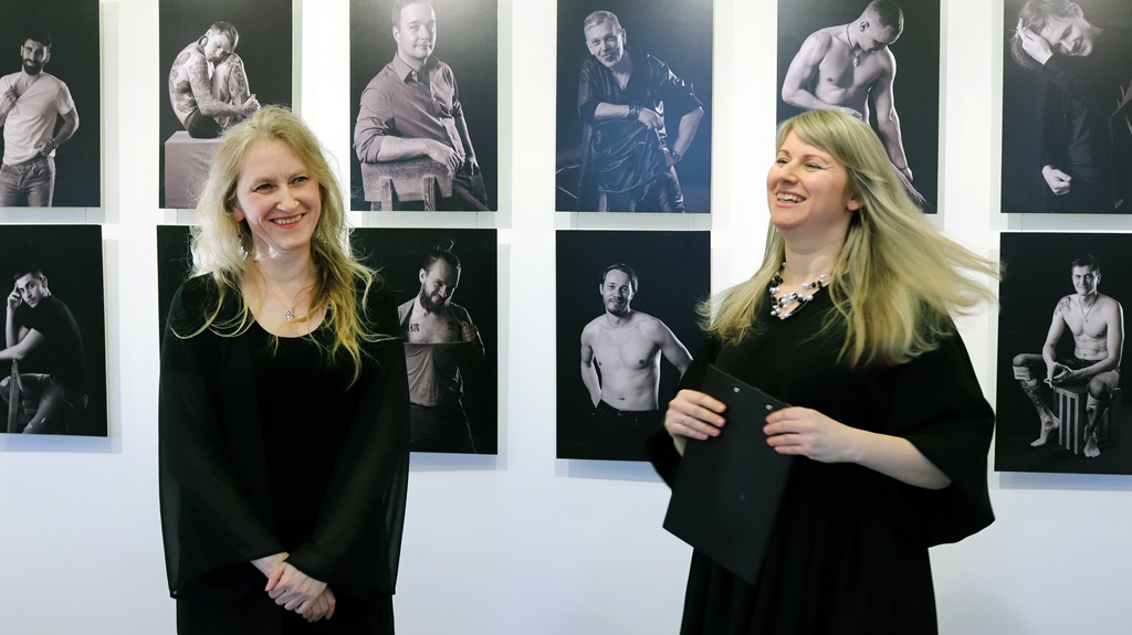 Divas sievietes stāv melnbaltu fotogrāfiju izstādes priekšā, iesaistījušās sarunā.