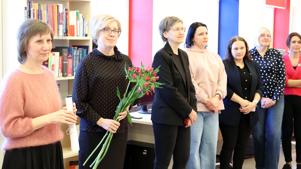 Sieviešu grupa stāv kopā grāmatu plaukta priekšā, kas pilns ar dažādām grāmatām.