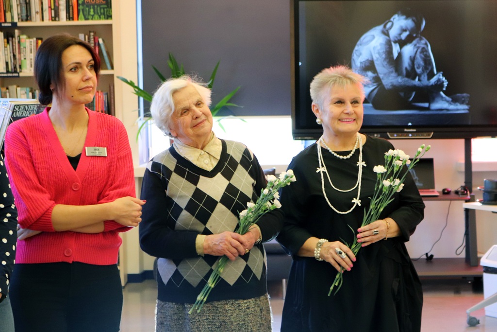 Trīs sievietes tur ziedus televizora ekrāna priekšā, smaida un kopā svin īpašu notikumu.