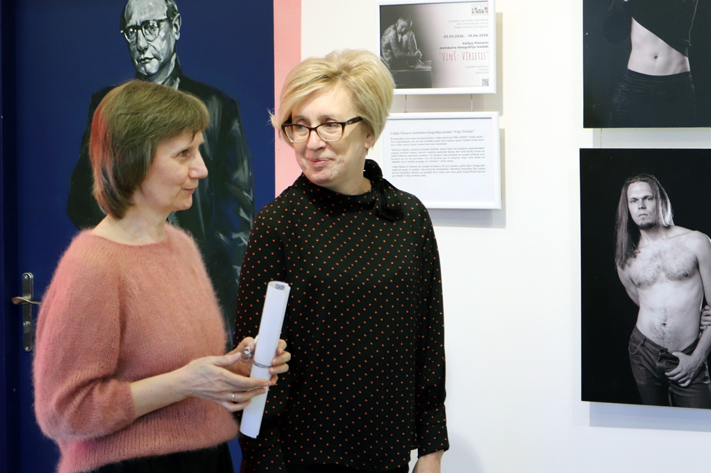 Divas sievietes stāv blakus, iesaistījušās sarunā mākslas izstādē, dažādu mākslas darbu ieskautas.