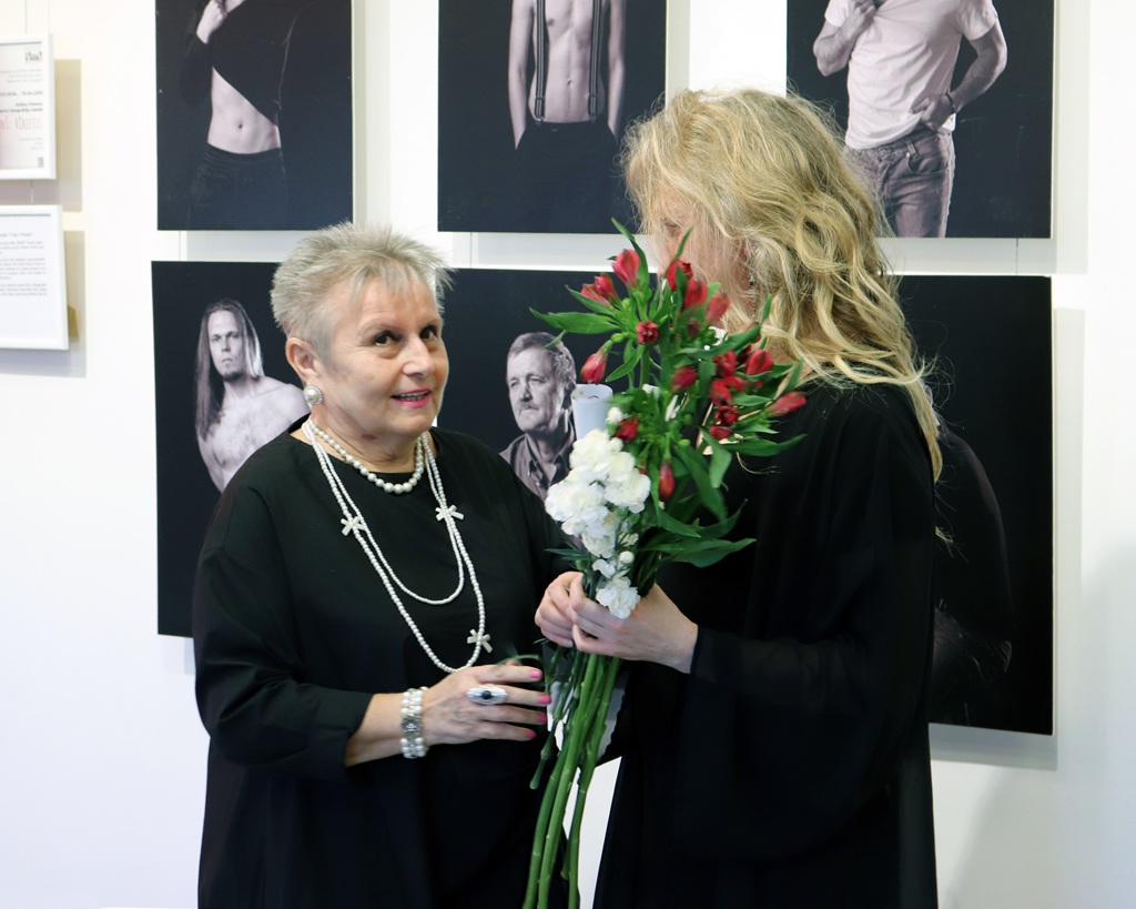 Sieviete tur ziedu pušķi, kamēr cita sieviete uz viņu skatās ar siltu sejas izteiksmi.