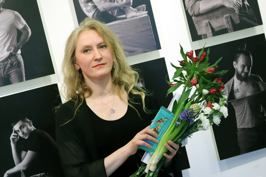 Sieviete tur ziedu pušķi ierāmētu gleznu kolāžas priekšā pie sienas.