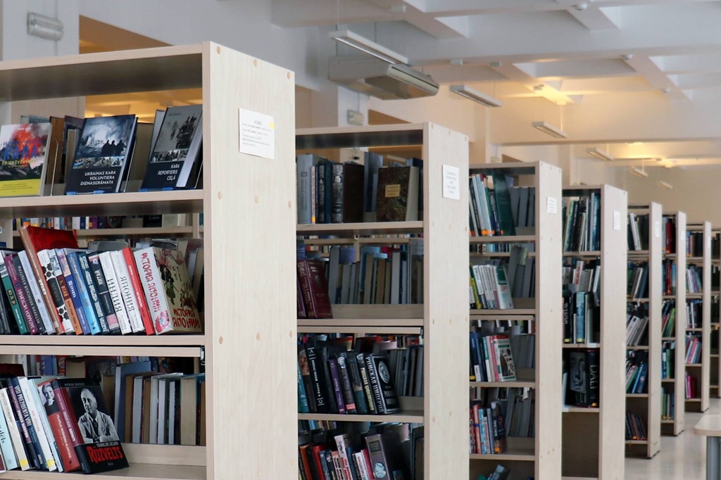 Spilgti apgaismotā bibliotēkā ir grāmatu plaukti, kas piepildīti ar dažādām grāmatām. Vide šķiet sakārtota un mierīga ar maigu griestu apgaismojumu.