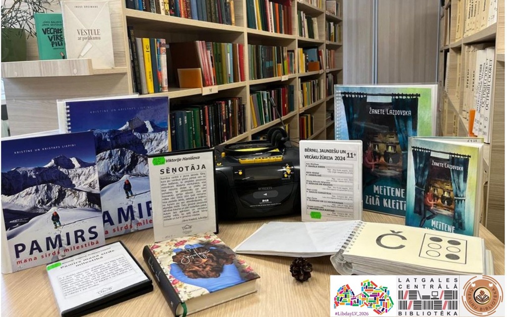 Bibliotēkas galdā ir izvietotas dažādas grāmatas krāsainos vākos, Braila raksta grāmata un audio atskaņotājs. Fonā redzami ar grāmatām piepildīti grāmatu plaukti.