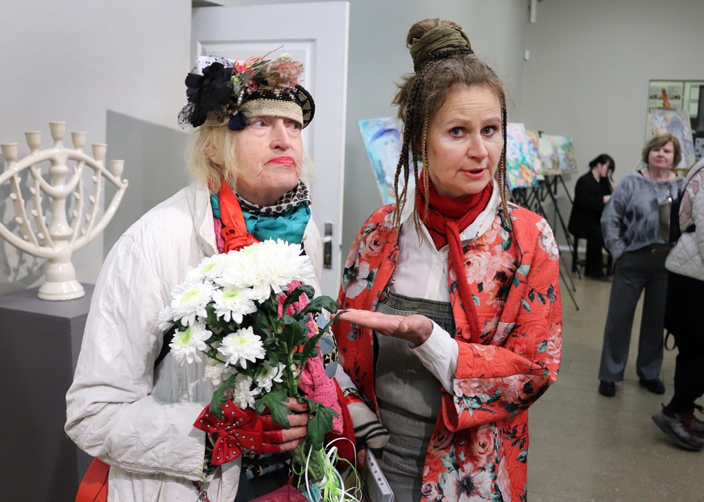 Divas sievietes krāsainos tērpos stāv mākslas galerijā; viena tur baltus ziedus, otra dzīvīgi žestikulē. Fonā redzamas gleznas un skatītāji.