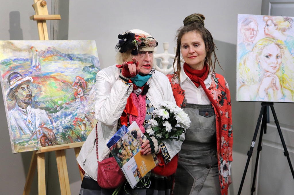 Divas sievietes stāv starp divām krāsainām gleznām, kas novietotas uz molbertiem. Sieviete kreisajā pusē ar ziedu raksta cepuri galvā un ziediem rokās dzīvīgi žestikulējas. Abas izskatās dzīvespriecīgas un iesaistītas.