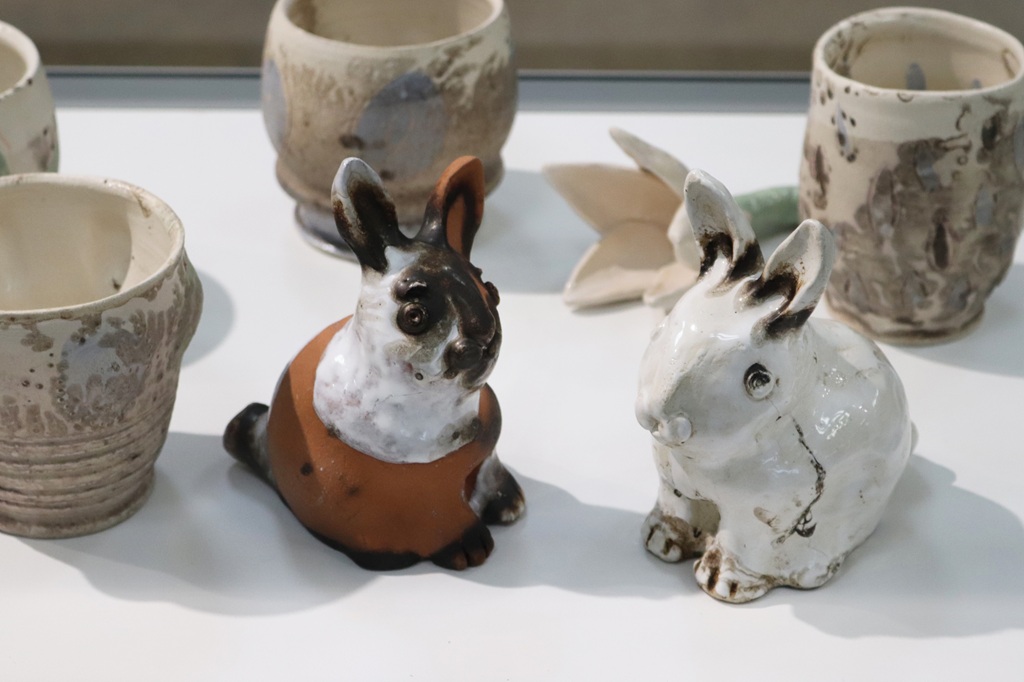 Divas keramikas trušu skulptūras, viena brūna un balta, otra netīri balta, atrodas starp rustikālām keramikas krūzēm uz baltas virsmas, radot rotaļīgu ainu.