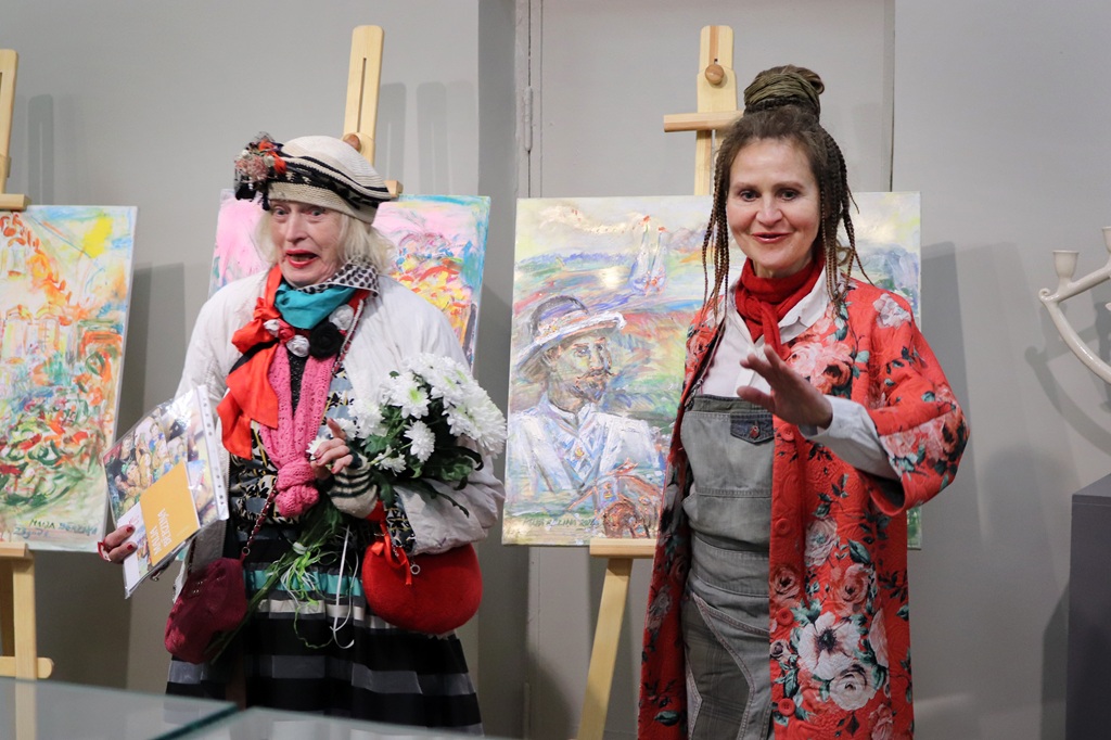Divas sievietes stāv krāsainu gleznu priekšā, kas novietotas uz molbertiem. Kreisā sieviete tur ziedus un papīrus, ģērbusies košā tērpā. Labā sieviete ar rotaļīgu sejas izteiksmi ir ģērbusies sarkanā ziedu raksta jakā. Abas izskatās dzīvespriecīgas mākslas izstādē.