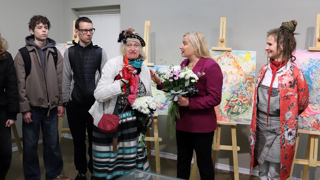 Dažāda veida cilvēku grupa stāv pie mākslas izstādes. Divas sievietes krāsainos tērpos tur rokās ziedu pušķus un sirsnīgi smaida, kamēr citas vēro. Noskaņojums ir svinīgs un dzīvespriecīgs.