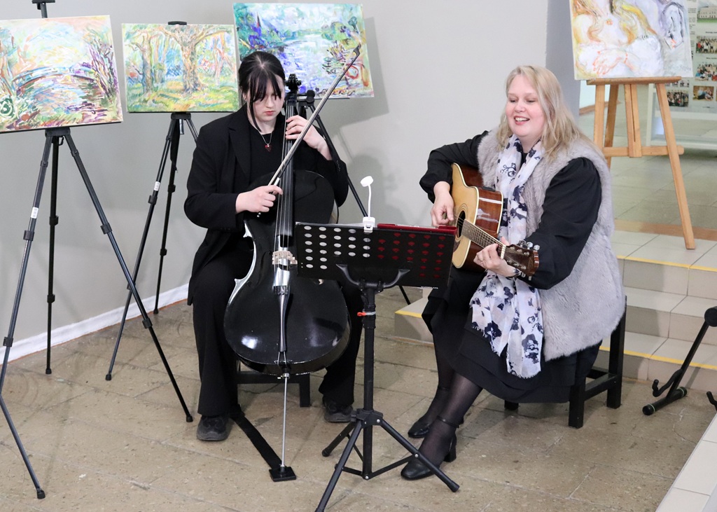 Divas sievietes uzstājas mākslas galerijā. Viena spēlē čellu, otra smaida, spēlējot ģitāru. Fonā uz molbertiem izvietotas krāsainas gleznas.