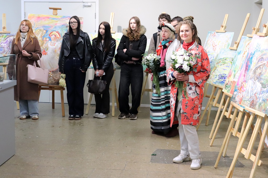 Cilvēku grupa stāv mākslas galerijā, vērojot krāsainas gleznas uz molbertiem. Sieviete ziedu raksta mētelī tur rokās pušķi, radot svētku noskaņu.