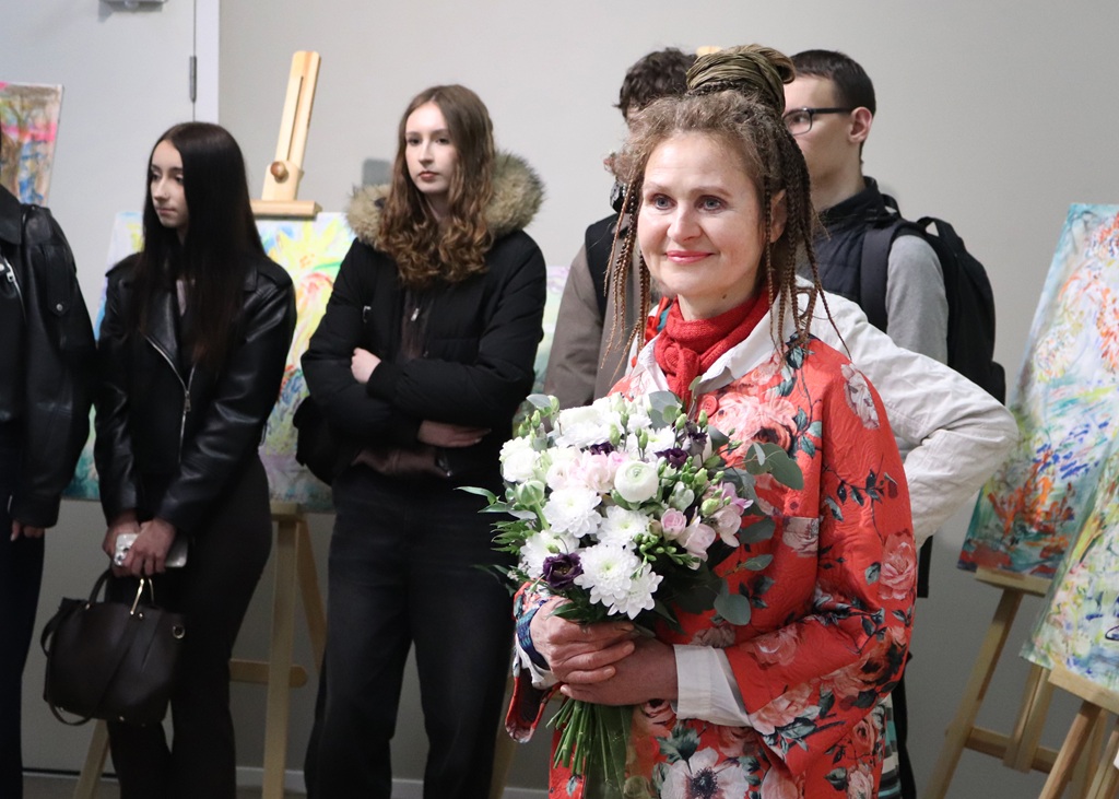 Sieviete sarkanā ziedu kleitā, turot rokās baltu un rozā ziedu pušķi, smaidot stāv mākslas galerijas priekšā, kuras fonā redzami vairāki cilvēki.