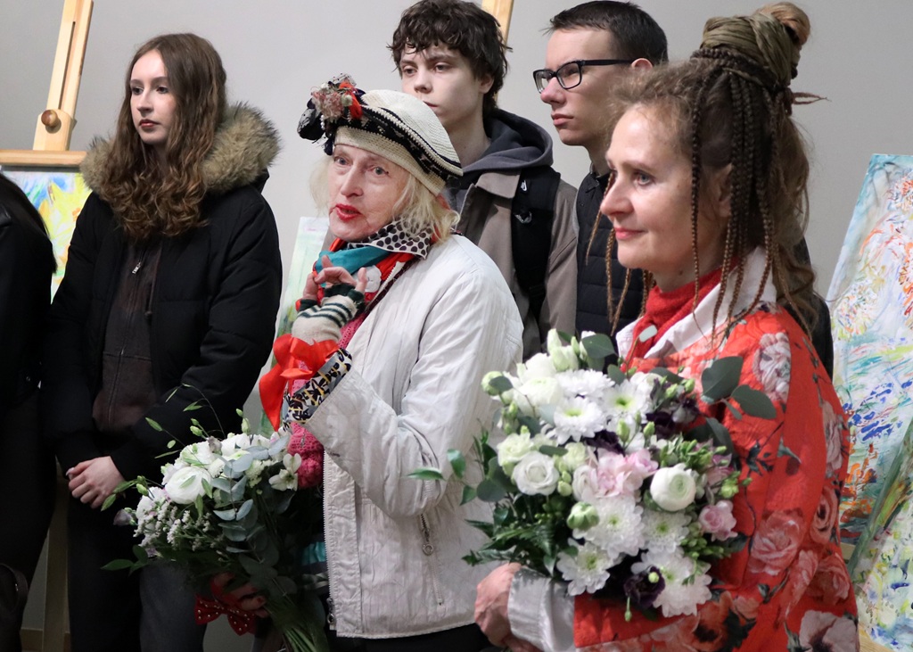 Divas sievietes tur rokās ziedu pušķus, stāv mākslas galerijā. Sieviete kreisajā pusē ir ģērbusies baltā jakā un cepurē, bet sieviete labajā pusē – sarkanā. Citi apmeklētāji, tostarp jauns vīrietis un sieviete, vēro notiekošo fonā. Atmosfēra ir dzīvīga un svinīga.