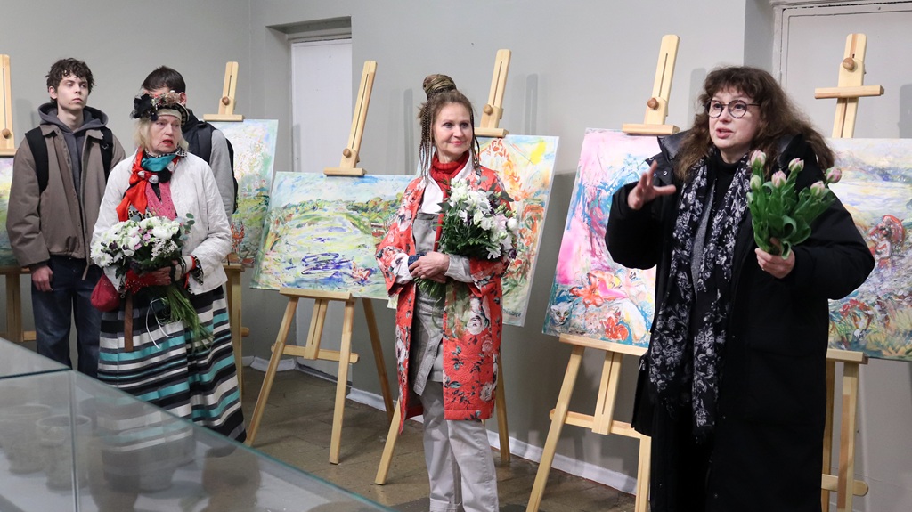 Trīs sievietes tur pušķus krāsainu gleznu priekšā, kas novietotas uz molbertiem, un abām pusēm atrodas divi vīrieši. Galerijas vidē valda svinīga un mākslinieciska noskaņa.