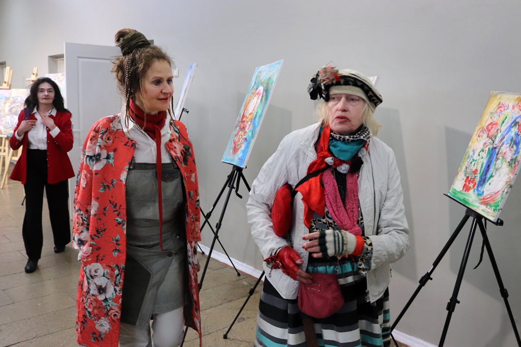 Divas sievietes košos, mākslinieciskos tērpos stāv un aplūko krāsainas gleznas uz molbertiem. Viņas atrodas mākslas galerijā, izstarojot radošumu un interesi.