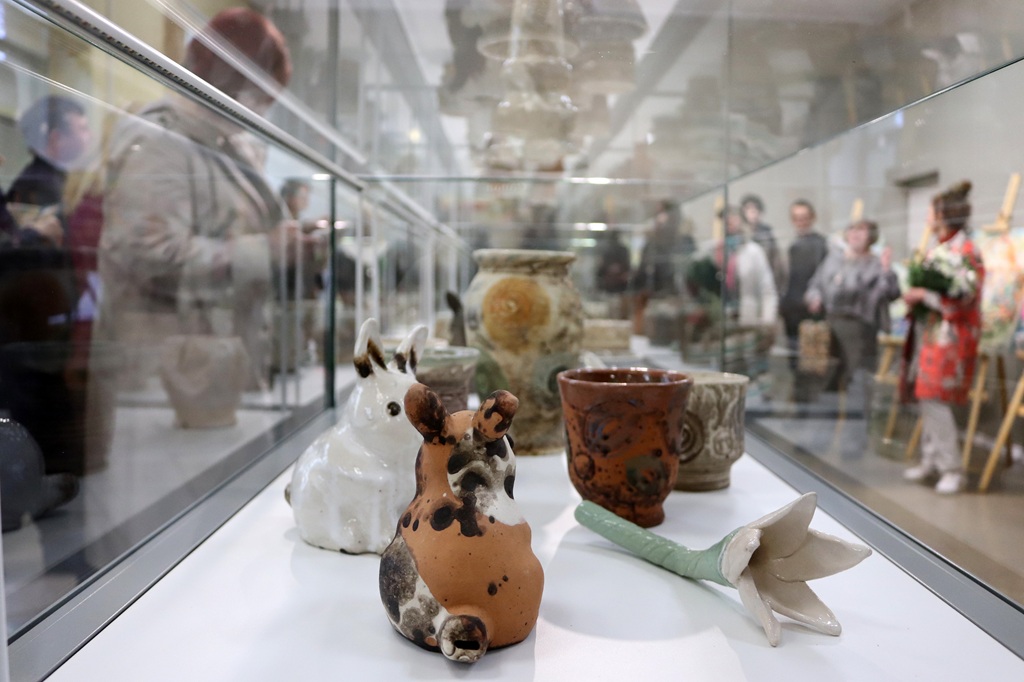 Keramikas figūru un vāzu izstāde stikla vitrīnā ar izplūdušiem apmeklētājiem fonā mākslas galerijā. Noskaņa ir dzīvīga un kulturāla.