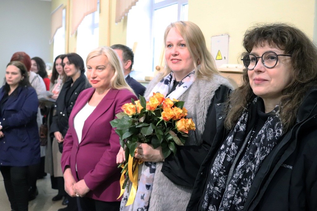 Sieviešu grupa stāv kopā labi apgaismotā telpā. Sieviete centrā tur oranžu rožu pušķi, radot dzīvespriecīgu atmosfēru.