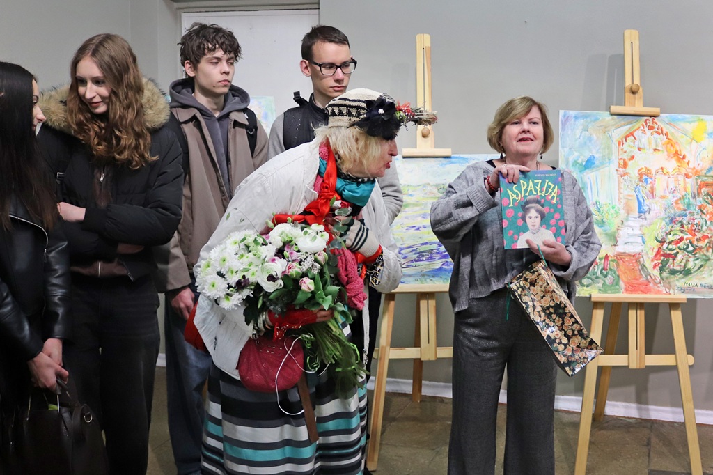 Cilvēku grupa stāv blakus krāsainām gleznām, kas novietotas uz molbertiem. Sieviete tur rokās grāmatu ar nosaukumu "Aspazija", bet cita, ar izrotātu cepuri, tur ziedus. Viņi izskatās priecīgi un iesaistīti.