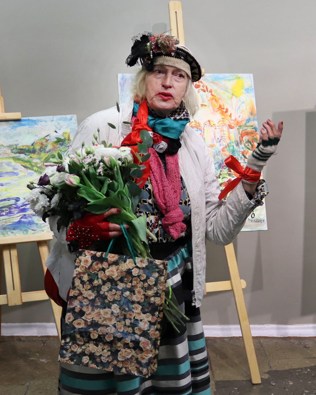 Izteiksmīgs vecāka gadagājuma cilvēks krāsainā, eklektiskā apģērbā tur rokās pušķi un dāvanu maisiņu. Fonā redzamas košas gleznas uz molbertiem.