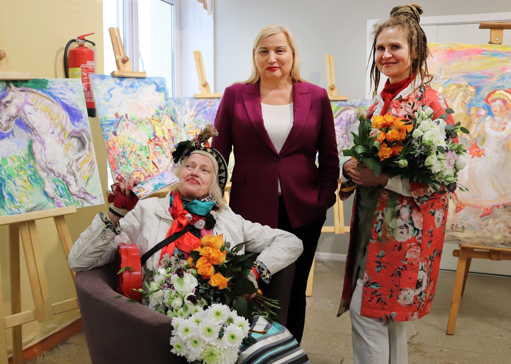 Trīs cilvēki atrodas mākslas galerijā, ko ieskauj košas gleznas uz molbertiem. Divi stāv, bet viens sēž krēslā, turot rokās pušķi. Noskaņojums ir svinīgs.