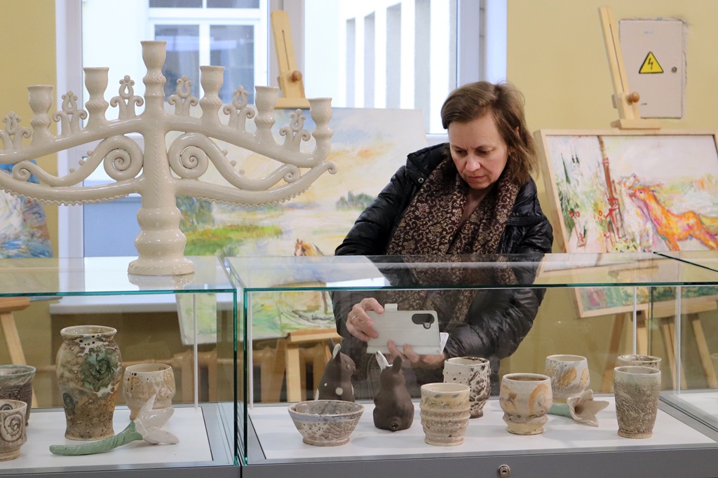 Sieviete fotografē keramikas mākslas darbus vitrīnā, ko ieskauj krāsainas gleznas uz molbertiem. Vide ir mierīga un radoša.