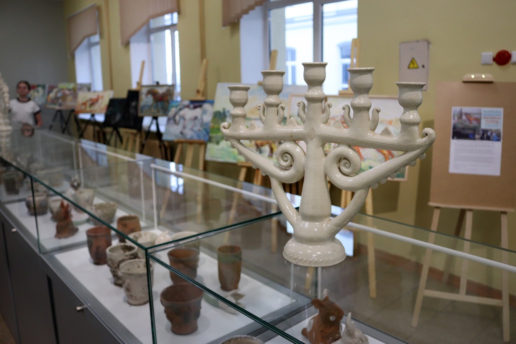 Balta keramikas menora ir izstādīta stikla vitrīnā līdzās dažādiem māla podiem. Fonā uz molbertiem novietotas gleznas rada māksliniecisku atmosfēru.