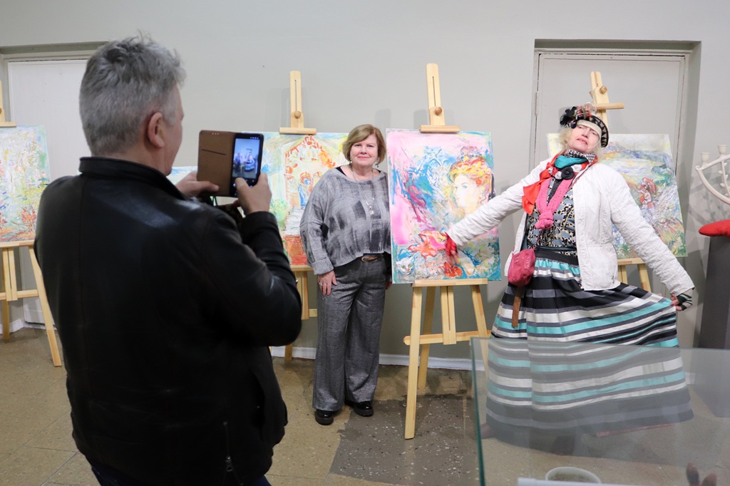 Vīrietis fotografē divas sievietes mākslas izstādē. Viena sieviete dinamiski pozē krāsainā apģērbā, bet otra mierīgi stāv blakus košām gleznām.