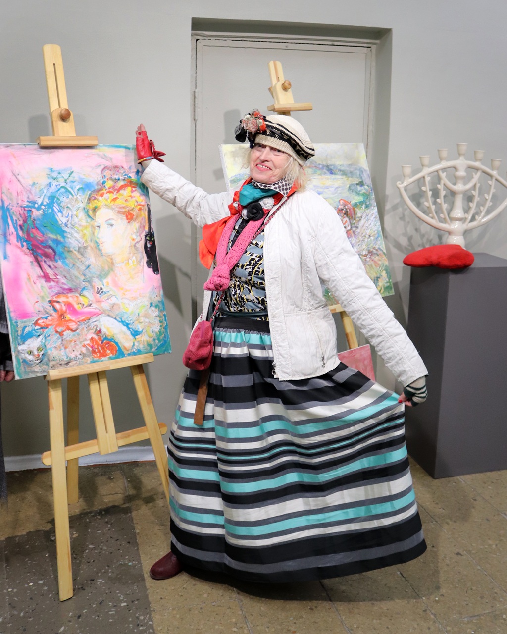 Cilvēks košā apģērbā priecīgi pozē blakus krāsainām abstraktām gleznām uz molbertiem. Vide ir mākslinieciska, izstaro dzīvīgu un radošu atmosfēru.