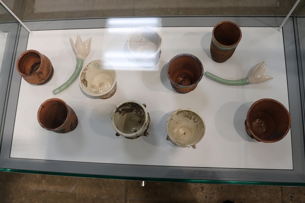 Keramikas vitrīna ar dažādiem brūniem un baltiem podiem, dažiem redzami ziedu kāti. Kārtīgi sakārtoti, mākslinieciskā un mierīgā noskaņā.