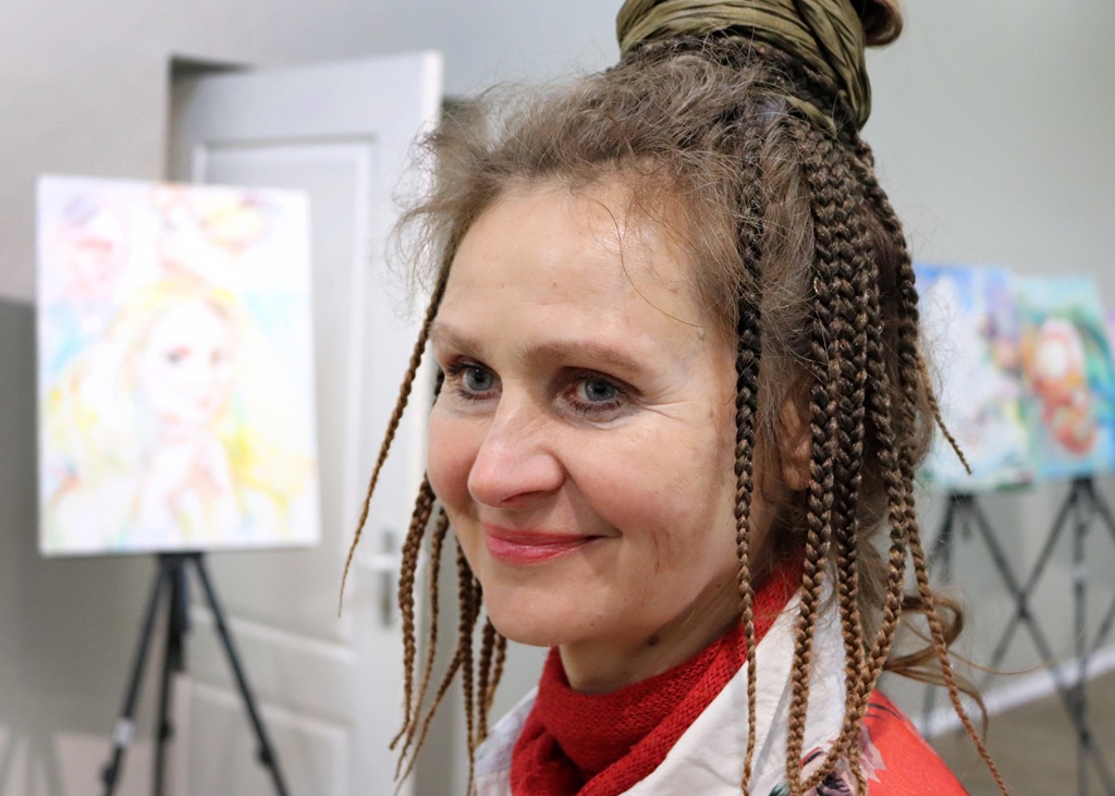 Smaidoša sieviete ar pītiem matiem un copi, sarkanā šallē, stāv mākslas galerijā. Fonā redzamas izplūdušas gleznas uz molbertiem.