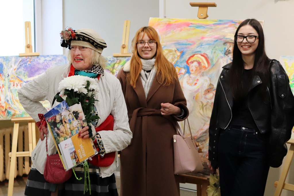 Trīs sievietes smaida mākslas galerijā. Sieviete kreisajā pusē tur ziedus un brošūras. Fonā uz molbertiem izvietotas krāsainas gleznas.
