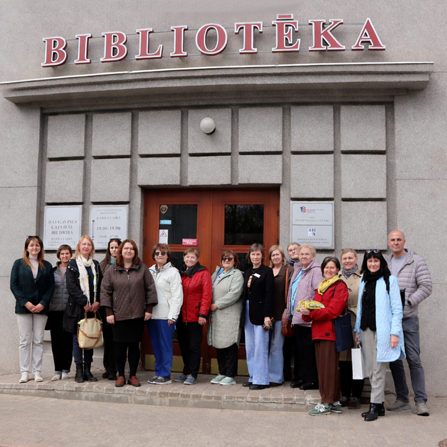 Cilvēku grupa smaidoši stāv ēkas priekšā, virs kuras ieejas ir uzraksts "Bibliotēka". Aina rada biedriskuma sajūtu.
