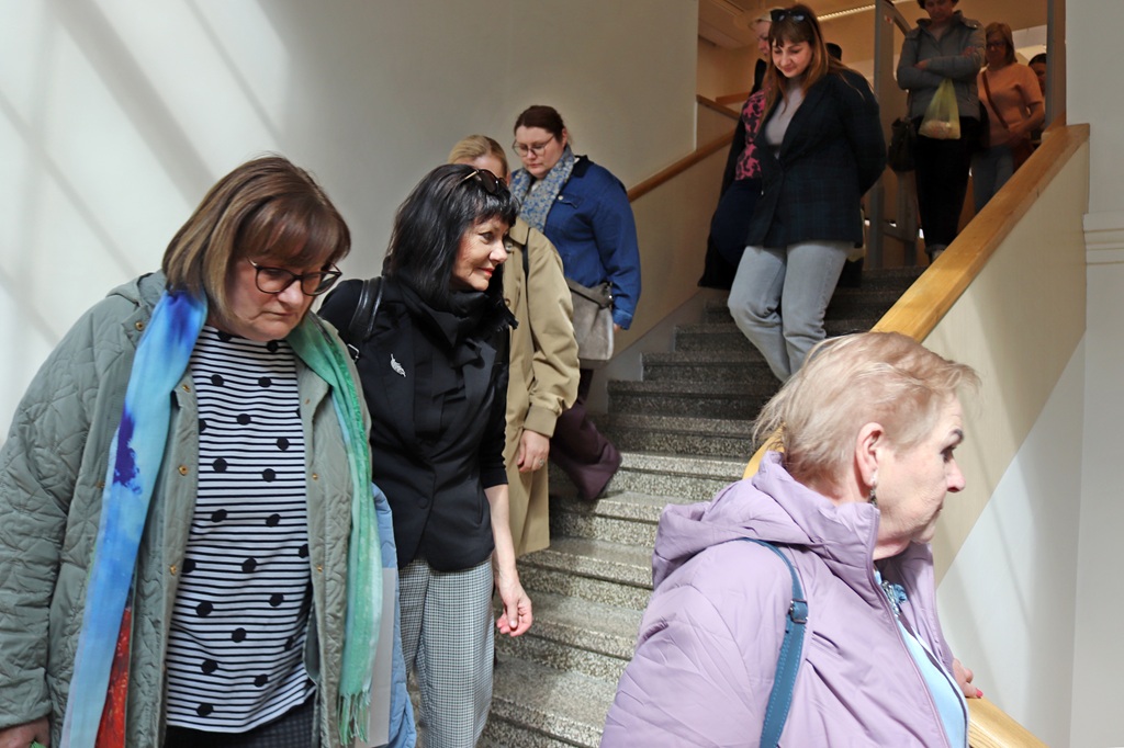 Cilvēku grupa, galvenokārt sievietes, kāpj lejup pa kāpnēm labi apgaismotā ēkā. Viņi izskatās koncentrēti un iesaistīti, ģērbušies ikdienas apģērbā.