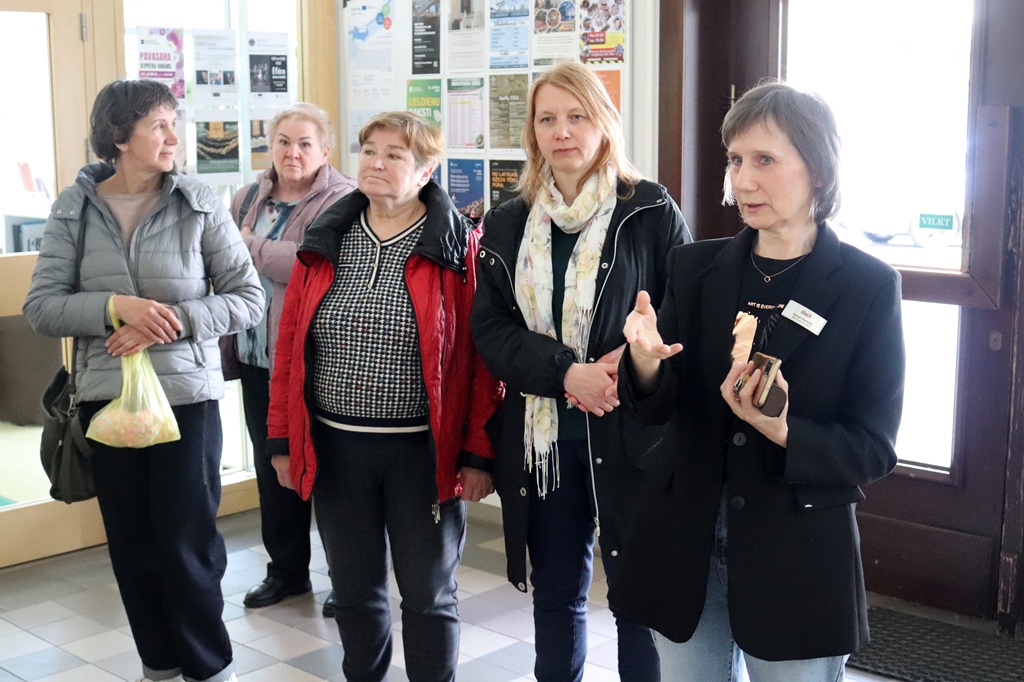 Piecas sievietes stāv iekštelpās, iesaistījušās diskusijā. Plakāti uz sienas aiz viņām liek domāt par kopienas vai publisku vidi. Atmosfēra ir uzmanīga un iesaistīta.