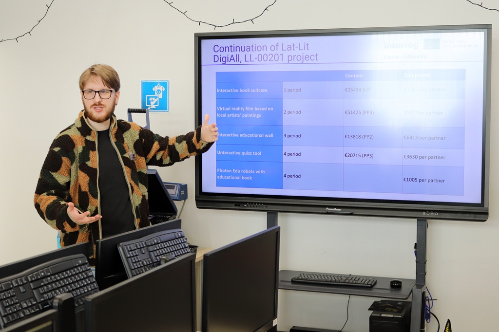 Rakstainā džemperī ģērbies cilvēks datorlaboratorijā uzstājas ar zīmi, norādot uz ekrānu, kurā redzams projekta turpināšanas plāns. Tonis ir informatīvs.