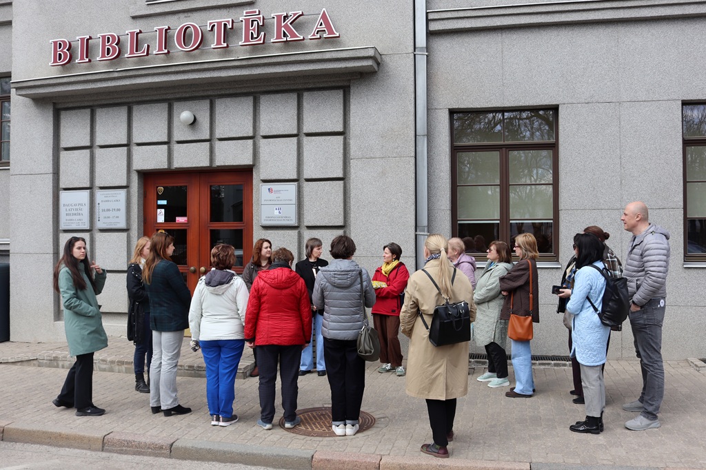 Cilvēku grupa stāv pie ēkas ar uzrakstu "Bibliotēka", kas liek domāt, ka tā ir bibliotēka. Šķiet, ka viņi ir iesaistījušies sarunā, iespējams, ekskursijā vai diskusijā.