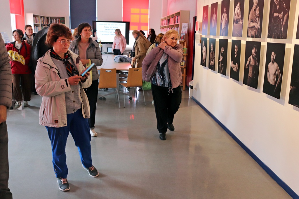 Cilvēku grupa bibliotēkas galerijas sienā vēro melnbaltu portretu sēriju. Atmosfēra ir pārdomu pilna un koncentrēta.