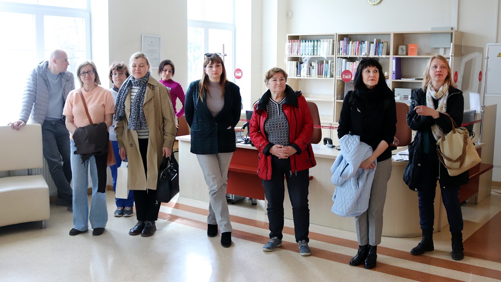 Deviņu cilvēku grupa stāv bibliotēkā, fonā grāmatu plaukti un logi. Atmosfēra ir neformāla, ar pārsvarā neitrālām sejas izteiksmēm.