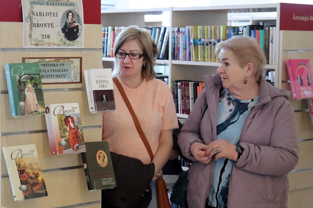 Divas sievietes stāv bibliotēkā un apspriež grāmatas, kas izliktas Šarlotes Brontē ekspozīcijā. Fonā plaukti ar krāsainām grāmatām.