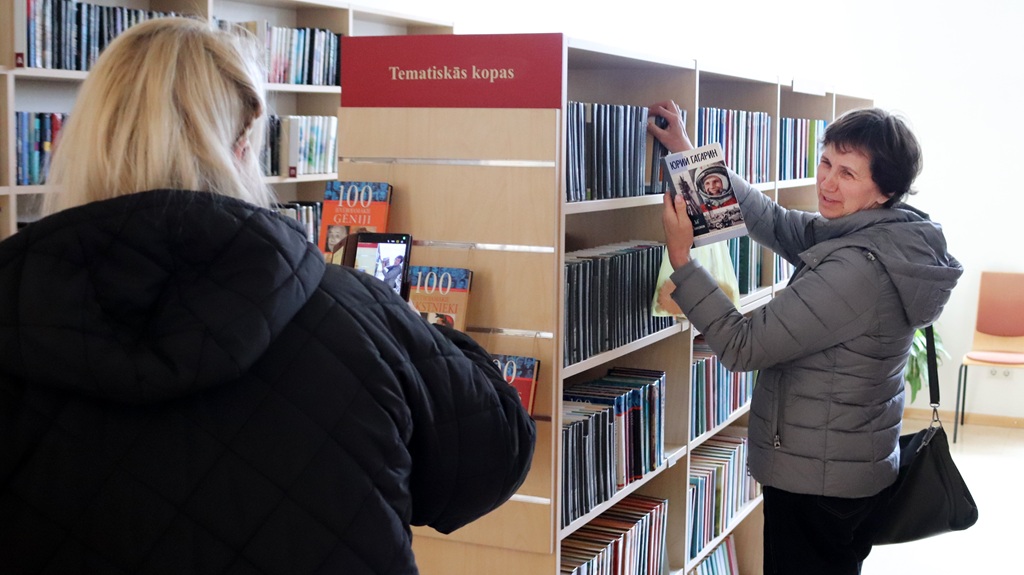 Sieviete pelēkā jakā tur grāmatu no bibliotēkas plaukta, kamēr cita persona fotografē. Plauktos ir glīti sakārtotas grāmatas.