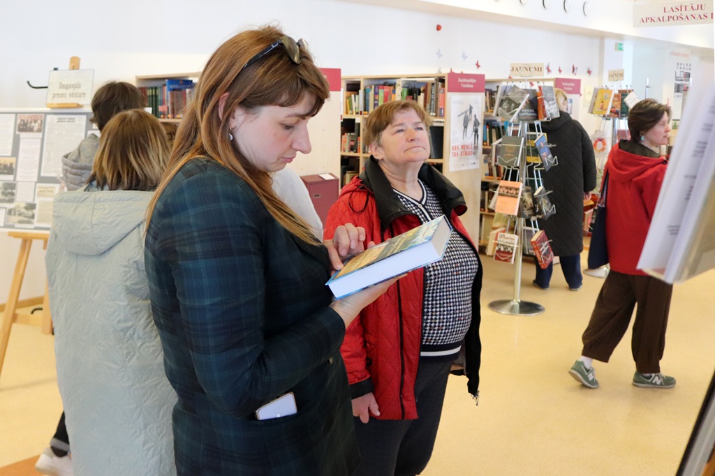 Sieviete rūtainā žaketē lasa grāmatu bibliotēkā. Citi apmeklētāji lasa grāmatas netālu, grāmatu plauktu un informatīvo displeju ieskauti. Mierīga atmosfēra.