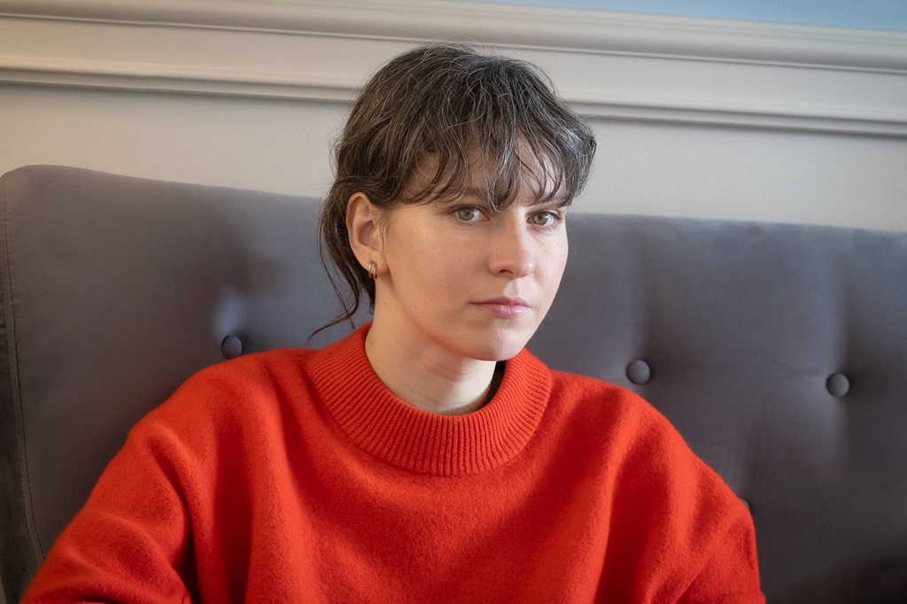 Cilvēks ar īsiem, tumšiem matiem sēž uz pelēka dīvāna, ģērbies spilgti sarkanā džemperī. Maigs apgaismojums un neitrāls fons liek viņam izskatīties domīgam.