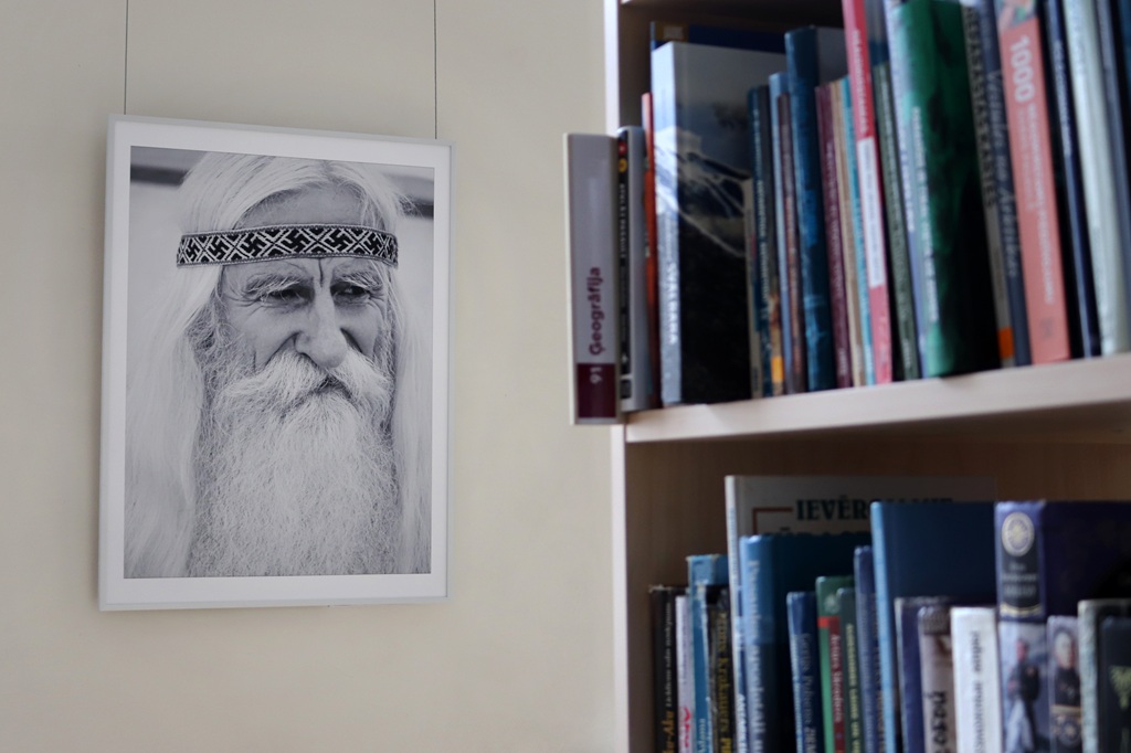 Melnbalts portrets, kurā attēlots vecāka gadagājuma cilvēks ar gariem, baltiem matiem un bārdu, kuram galvā rakstaina galvas saite. Fotogrāfija karājas blakus grāmatu plauktam, kas pilns ar krāsainām grāmatām.